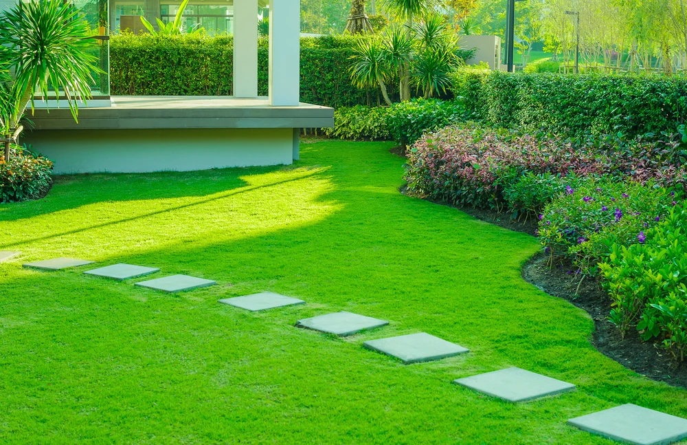 Vue aérienne d'un jardin paysager moderne avec terrasses en bois composite, mobilier d'extérieur blanc, pelouse synthétique ou naturelle parfaitement entretenue, bordures courbes en béton, massifs d'arbustes taillés en boules, végétation périphérique dense