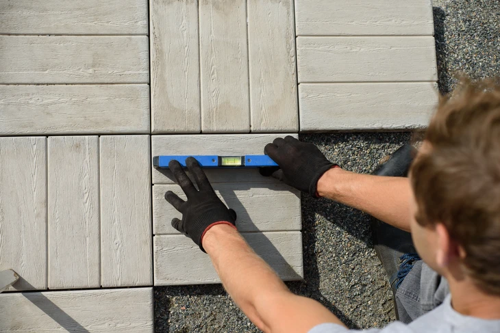 Ouvrier en gants noirs utilisant un niveau à bulle bleu sur des dalles de terrasse extérieure en béton imitation bois