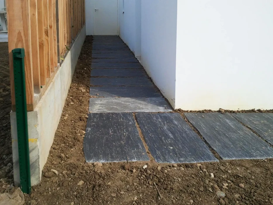 Allée extérieure entre un mur de maison enduit blanc et une clôture en bois sur muret béton