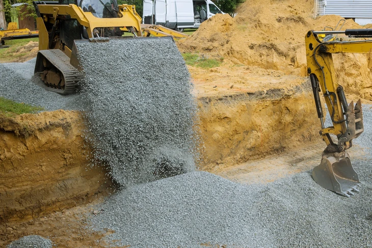 Chantier de construction extérieur avec pelleteuse jaune déversant du gravier gris sur terrain excavé